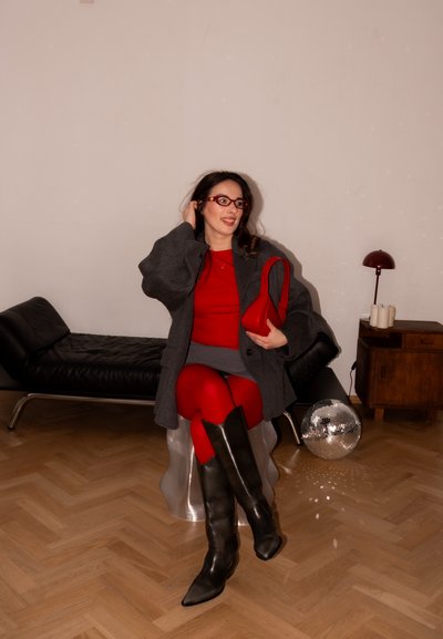 Mujer con vestido rojo, abrigo gris, botas negras y gafas rojas sentada en un taburete plateado, sosteniendo un bolso rojo en una habitación moderna.