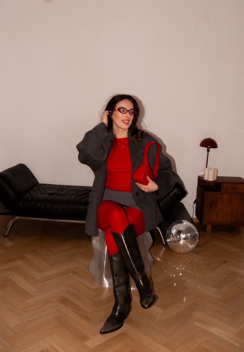 Femme en robe rouge, manteau gris, bottes noires et lunettes rouges assise sur un tabouret argenté, tenant un sac rouge dans une pièce moderne.