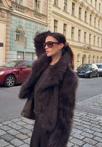 Jachetă din blană ecologică maro cu un guler larg, purtată peste un top închis la culoare. Ochelari de soare mari și păr împletit, stând pe o stradă pietruită.