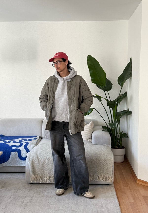 Hombre con gafas y gorra roja de pie en el salón, lleva una sudadera gris con capucha, chaqueta verde acolchada, vaqueros negros anchos, junto a un sofá gris y una planta alta y verde.