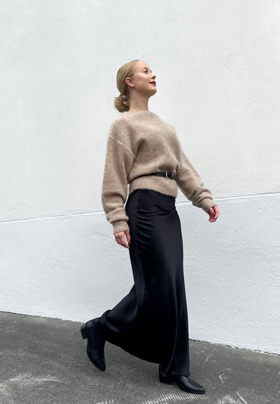 Suéter de punto beige, falda de satén negra, botas negras de tobillo. La modelo está de pie contra una pared blanca, caminando con un ligero giro en su postura.