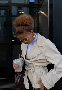 Kvinde med krøllet hår iført en beige trenchcoat og store øreringe, der holder en kold drik med et sugerør udenfor indgangen til en bygning.