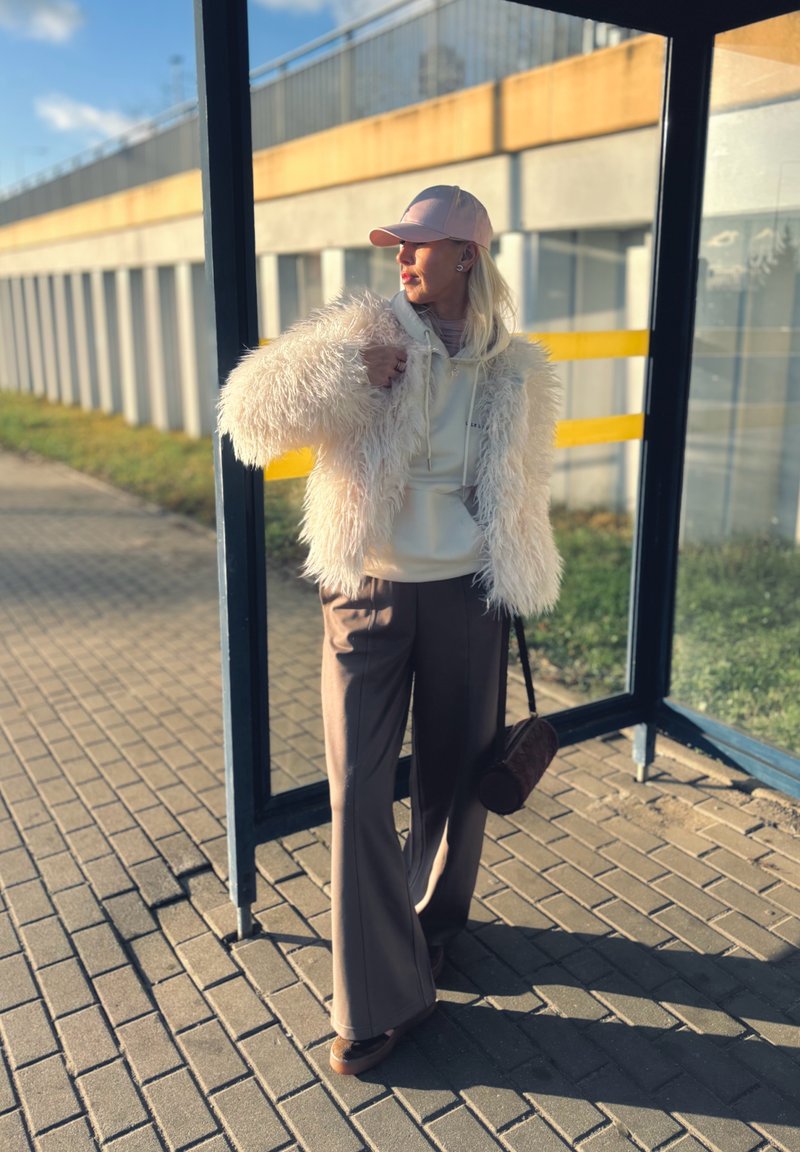 Apresenta um casaco branco fofo, um capuz leve, calças castanhas de perna larga e um boné rosa. Os acessórios incluem uma mala redonda castanha.