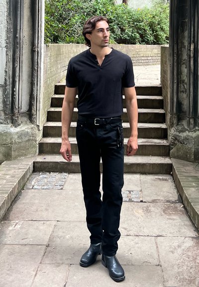 Camisa henley negra de manga corta, jeans negros de corte ajustado y botas negras hasta el tobillo, de pie en escalones de piedra con vegetación al fondo.