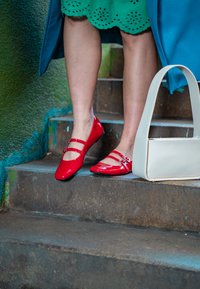 Κόκκινες παλτά δερμάτινες μπαλαρίνες με διπλές λωρίδες, τοποθετημένες πάνω σε πέτρινες σκάλες. Μια μαλακή λευκή τσάντα βρίσκεται κοντά, προσθέτοντας αντίθεση.