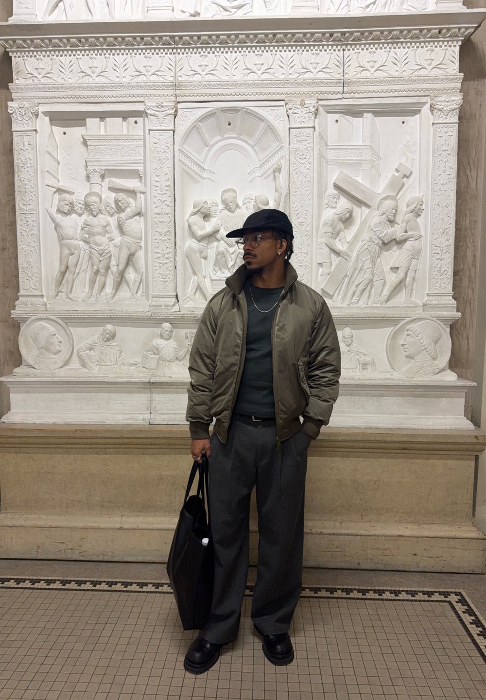 Homme portant une casquette, des lunettes, une veste verte et un pantalon gris tenant un sac noir, debout devant un relief religieux en pierre blanche détaillé.