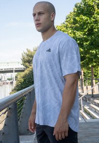 Hellgraues Sport-T-Shirt mit strukturiertem Muster, Rundhalsausschnitt, kurzen Ärmeln und einem kleinen schwarzen Logo auf der linken Brust.