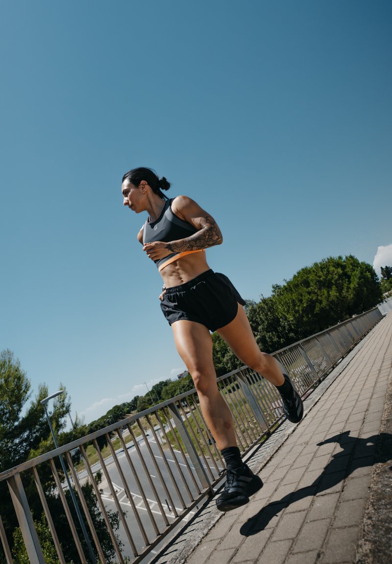Atletisk kvinna med tatueringar joggar på en upphöjd trottoar bredvid ett metallräcke under en klar blå himmel.