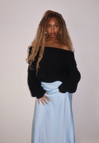 Sweater preto à off-the-shoulder, malha solta. Saia satin azul claro, textura suave, até ao tornozelo, mãos nos quadris, joias em camadas visíveis.