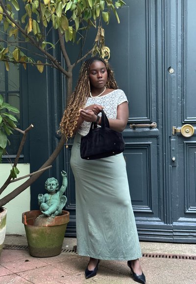 Bolso de ante negro, blusa de encaje blanca, falda verde menta y tacones negros. El fondo incluye una planta en maceta y una estatua decorativa.