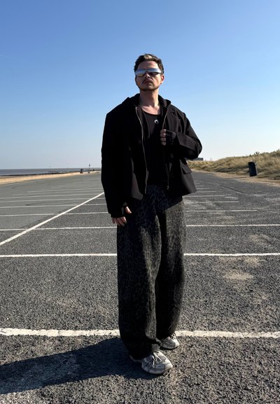 Chaqueta negra, camiseta de tirantes negra y pantalones sueltos de estampado de leopardo negro. Zapatillas grises. Entorno al aire libre con un cielo azul claro.