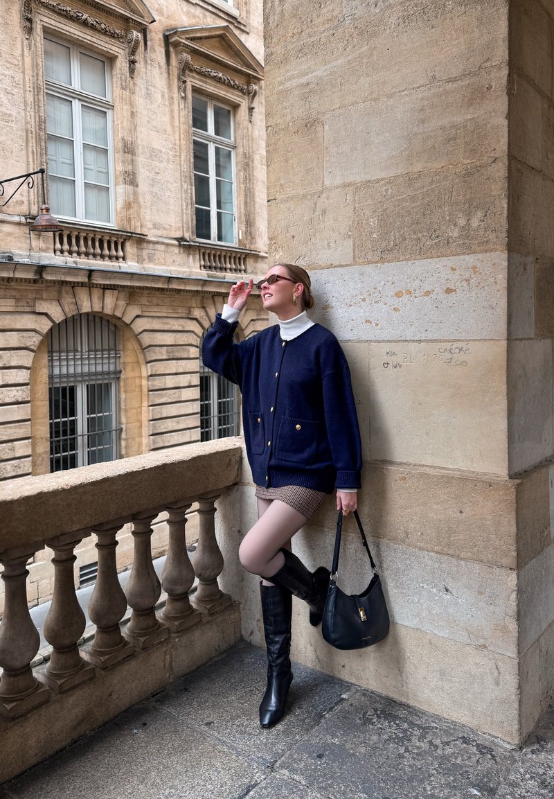 Femme en cardigan bleu marine et jupe à carreaux appuyée contre un mur en pierre sur un balcon, ajustant ses lunettes de soleil, tenant un sac à main noir.