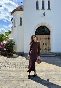 Top marrón de manga larga y falda midi a juego con una abertura lateral, combinados con zapatos negros tipo slip-on y un pequeño bolso marrón. Camino de piedras y edificio blanco de fondo.