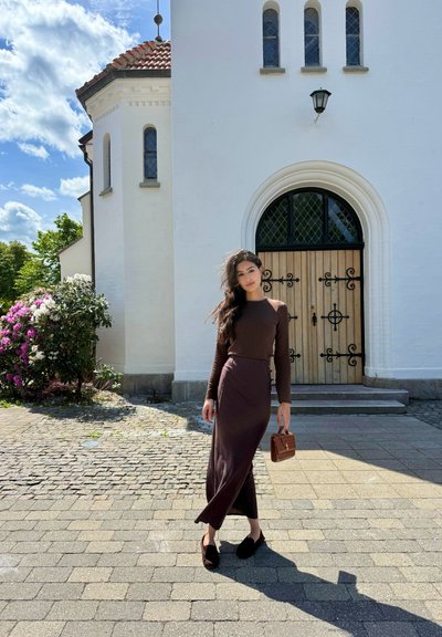 Top marrón de manga larga y falda midi a juego con una abertura lateral, combinados con zapatos negros tipo slip-on y un pequeño bolso marrón. Camino de piedras y edificio blanco de fondo.
