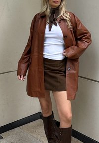 Bruin leren jasje met lange mouwen, knoopsluiting en een lichte glans, gecombineerd met een bruin suède minirok en donkerbruin kniehoog laarzen.