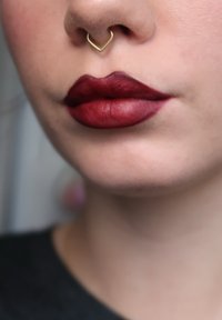 Close-up van volle lippen met donkere rode matte lippenstift en een gouden hartvormige neusring. De gladde textuur en gedefinieerde lipcontour zijn zichtbaar.
