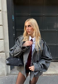 Cappotto grigio oversize con motivo a pied de poule, bottoni neri e maniche voluminose, abbinato a una camicia bianca, cravatta color borgogna e borsa nera.