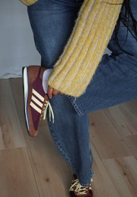 Des chaussures de sport marron et bordeaux avec des lacets jaunes, arborant trois bandes blanches ; portées avec un jean bleu et un pull jaune texturé.