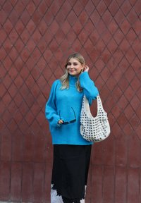 Turkusowy oversize'owy sweter z wysokim kołnierzem, zestawiony z czarną spódnicą zdobioną koronką. Trzyma okulary przeciwsłoneczne, niesie białą torebkę z ćwiekami.