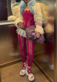 Personne portant une veste en fourrure par-dessus un body en dentelle rose à lacets et des leggings, des sandales plateformes blanches, tenant un sac violet, debout dans un ascenseur métallique.