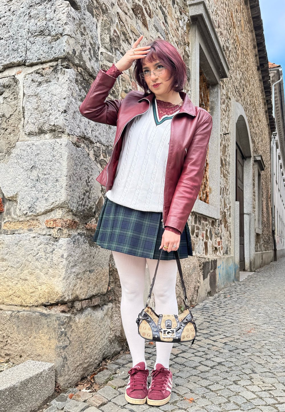 Burgundijska usnjena jakna, bela pletena majica, karirana krila, bele najlonke, burgundijske tenisice in torbica s potiskom. Ozadje iz kamnitih zidov.