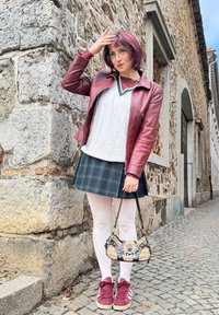 Burgundijska usnjena jakna, bela pletena majica, karirana krila, bele najlonke, burgundijske tenisice in torbica s potiskom. Ozadje iz kamnitih zidov.