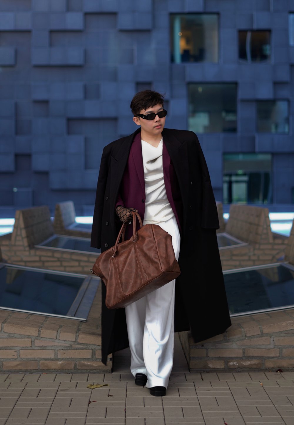 Καφέ δερμάτινη τσάντα duffle, μαύρο μακρύ παλτό, λευκά παντελόνια και μοβ μπλουζάκι; συνδυασμένα με μαύρα γυαλιά ηλίου και δαντελένια γάντια. Σύγχρονο φόντο.