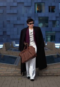 Καφέ δερμάτινη τσάντα duffle, μαύρο μακρύ παλτό, λευκά παντελόνια και μοβ μπλουζάκι; συνδυασμένα με μαύρα γυαλιά ηλίου και δαντελένια γάντια. Σύγχρονο φόντο.