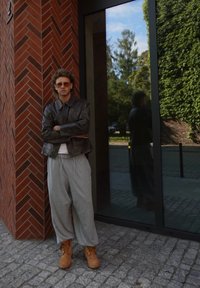 En person i en svart skinnjacka, grå joggebukser og beige støvler står mot en murvegg, reflektert i et glasspanel.