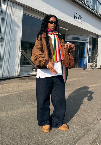 Chaqueta de cuero marrón sobre una camisa blanca, jeans de pierna ancha de color azul oscuro, bufanda de rayas coloridas y bolso marrón. Gafas de sol y calzado casual.