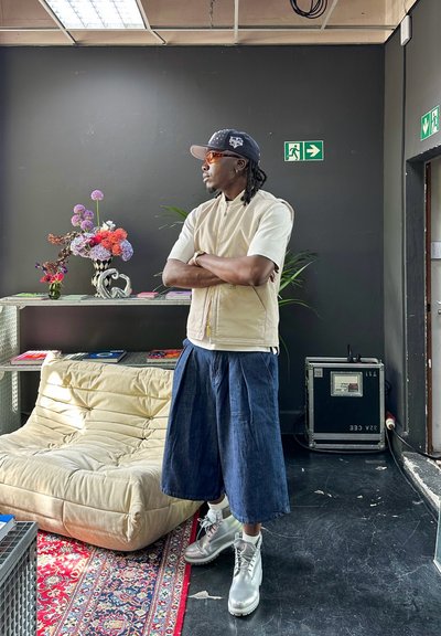 Chaleco beige con cremallera sobre una camisa blanca, pantalones anchos azules y botas grises. Entorno interior con flores, estantería y paredes negras.