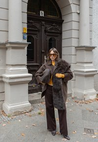 Barna gyapjúkabát műszőrmével, mustársárga pulóver, barna nadrág és szürke kézitáska. Átlátszó napszemüveg és bőr magassarkú csizma. Városi környezet.