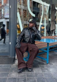 Homme en veste en cuir noire, gants et casquette assis sur un banc bleu devant un café, regardant sur le côté avec des bâtiments reflétés dans le verre derrière lui.