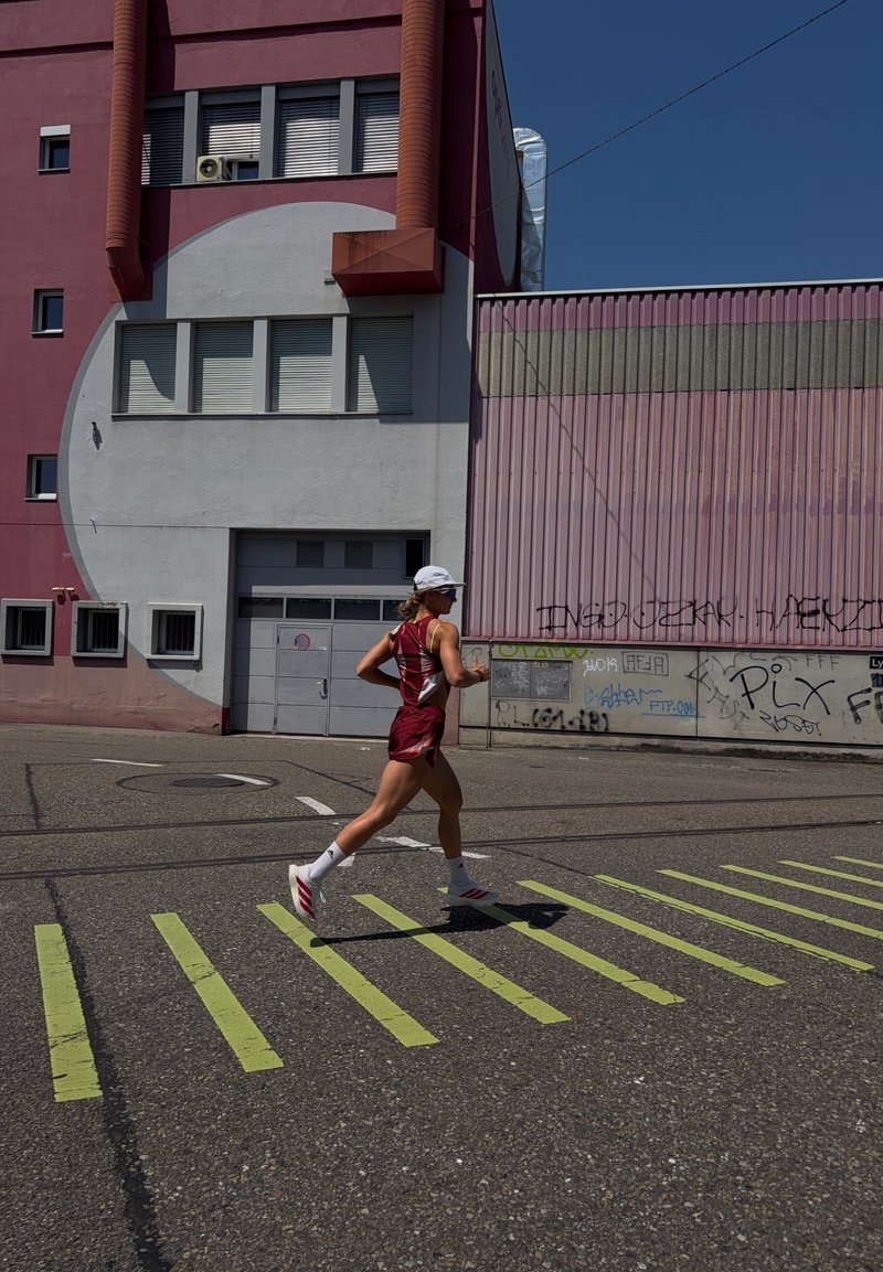 Coureur portant une tenue de sport rouge et une casquette blanche, courant à travers une rue avec des marquages routiers jaunes devant un bâtiment rose et gris couvert de graffitis.
