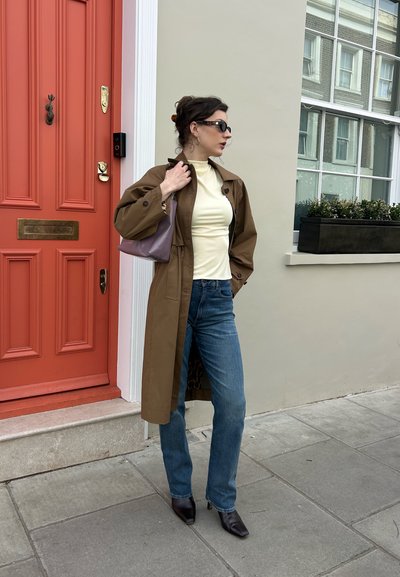 Mujer que lleva un abrigo marrón, top crema, vaqueros azules, botas negras y gafas de sol, de pie sobre la acera cerca de una puerta roja y una ventana con macetero.