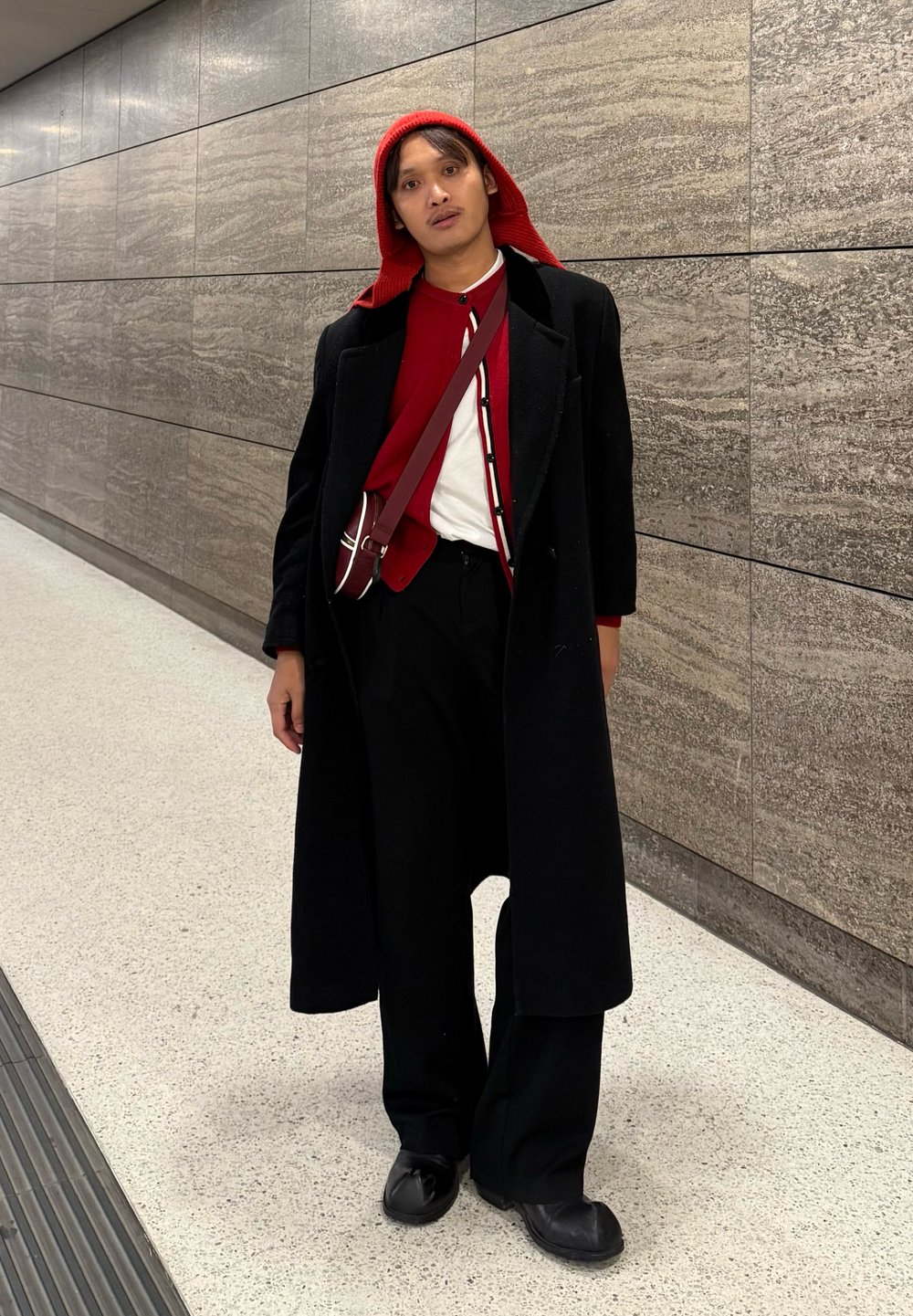 Long manteau noir porté sur un haut rouge, pantalon noir à jambes larges et capuche rouge. Un petit sac avec des accents contrastants est porté en bandoulière.