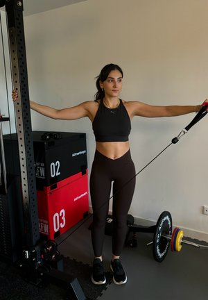 Entrenamiento de resistencia con cables, con una persona usando un top corto negro y leggings marrones. Cerca hay cajas de ejercicio rojas y negras, con pesas en el suelo.