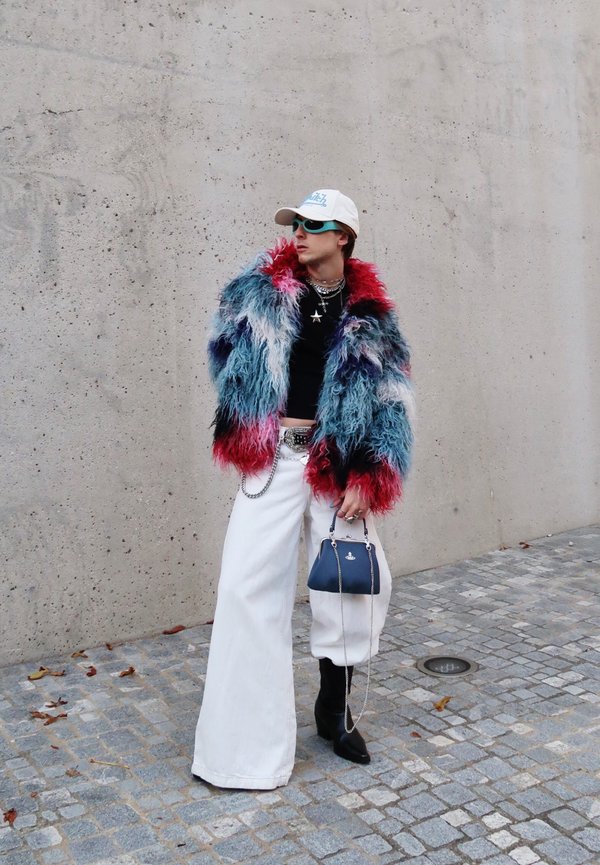 Person iført en flerfarvet fluffy jakke, hvide vidde bukser, sorte støvler, hvid kasket, turkise solbriller og holder en lille marineblå håndtaske på en brostensbelagt gade.