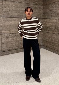 Pull tricot rayé marron et blanc, pantalon noir à jambes larges, et chaussures marron foncé. Fond uni avec mur texturé.