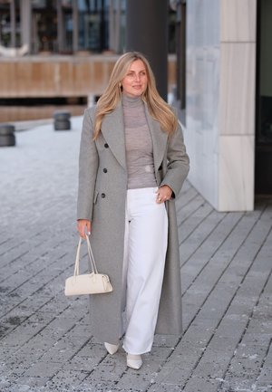 Mujer caminando al aire libre con un abrigo largo gris, un suéter de cuello alto gris, pantalones blancos, tacones blancos y llevando un pequeño bolso beige.