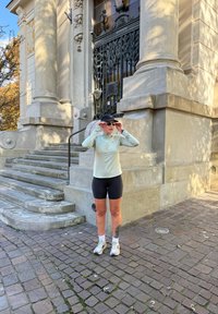 Tenue de sport comprenant un haut à manches longues vert clair et un short de cyclisme noir. Accessoires : une casquette noire et des lunettes de soleil. Fond en pavé de briques.