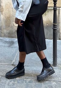 Černé kožené slip-on boty s robustními podrážkami a texturovaným vzorkem, nošené s černými ponožkami a sukní ke kolenům v černé barvě.