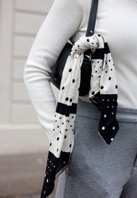 Chemise blanche à manches longues côtelées, pantalon gris, sac à bandoulière noir avec un foulard noir et blanc à pois attaché à la sangle.