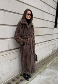 Manteau en fausse fourrure marron avec ceinture, col oversize et poches. Porté sur un col roulé noir avec des gants en cuir noir et des bottines.