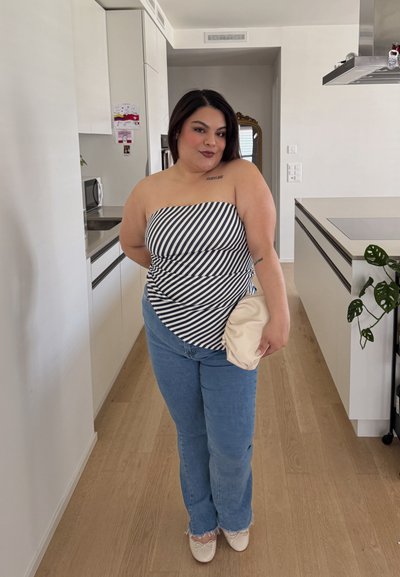 Top sin tirantes a rayas y jeans de un azul claro con dobladillos deshilachados. Sosteniendo un clutch de color crema; de pie en una cocina moderna con un suelo de madera.
