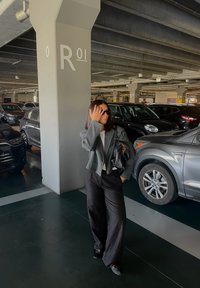 Ženska v sončnih očalih, sivo bluzo in črnimi hlačami hodi po večnadstropnem parkirišču v bližini parkiranih avtomobilov in betonskega stebra označenega z R01.