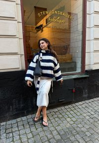 Gestreifter Pullover in Marineblau und Weiß, kombiniert mit einem weißen Rock und Flip-Flops, steht in der Nähe einer Bäckerei mit gefliesten Fenstern.