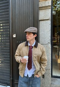 Kerge pruun zip-up jakk tumeda kraega, valge särk, triibuline lips, lame kübar ja valge kohvitass käes. Kerge denimist teksad.