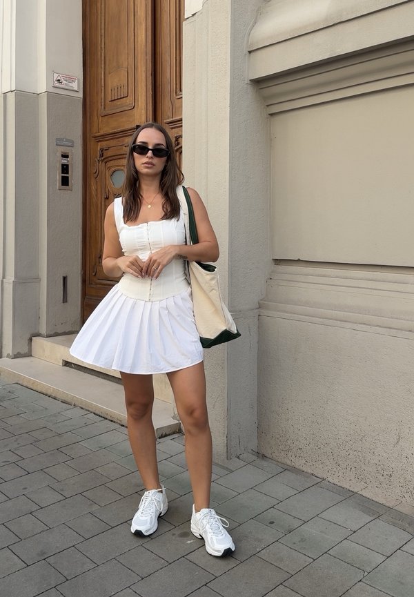 Žena v bielych šatách a teniskách so slnečnými okuliarmi a taškou stojí na chodníku pred drevenými dverami.
