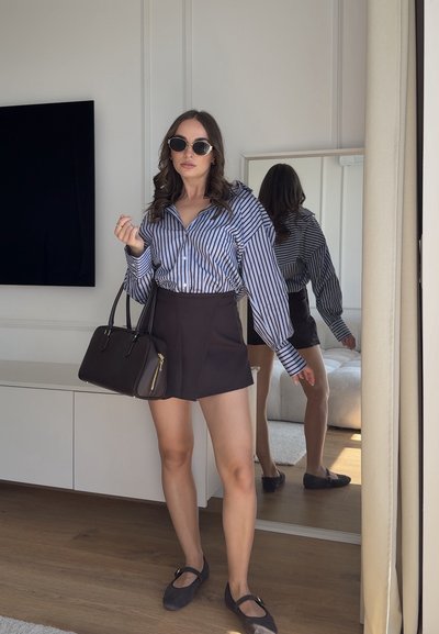 Mujer con gafas de sol, blusa de rayas, falda negra y zapatos planos, sosteniendo un bolso negro, de pie en el interior cerca de un espejo de cuerpo entero.